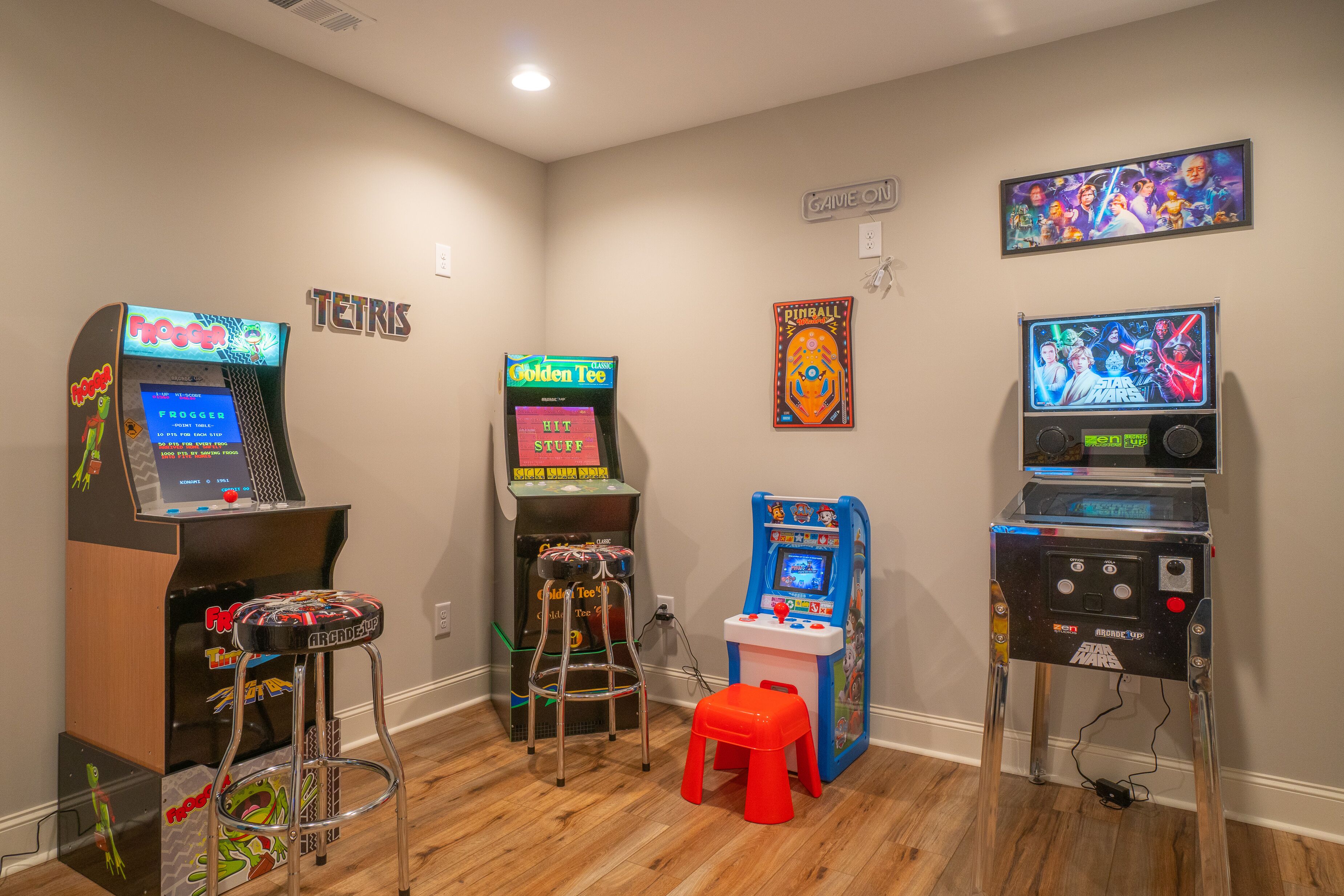 A room with four arcade machines, a small red stool, and a Star Wars themed poster on the wall.