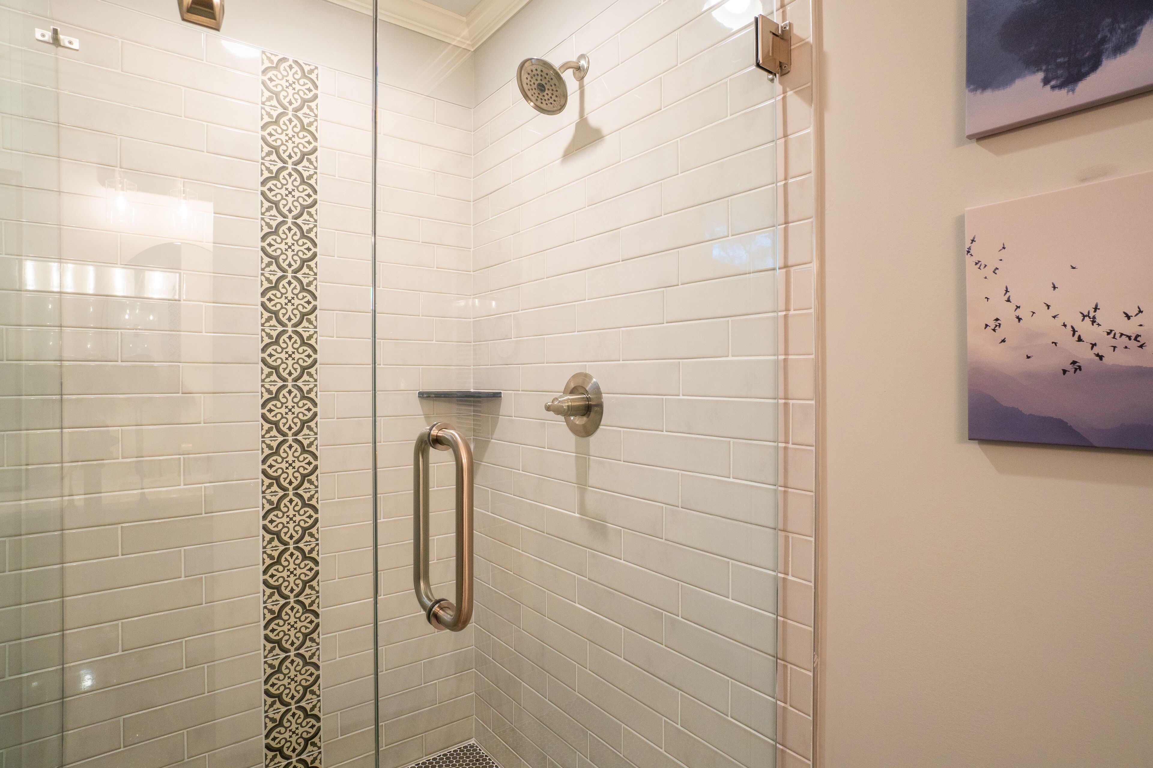 A shower with white tiles and a decorative border, a handheld showerhead, and a glass door.