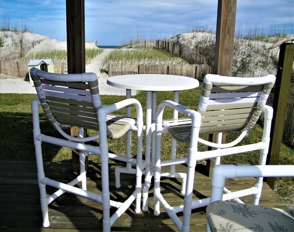 A deck with white plastic chairs and a table, overlooking a sandy beach and dunes.