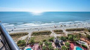 Blick auf einen Strand von einem Balkon mit Poolbereich, Liegestühlen und Sonnenschirmen.
