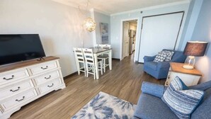 A living room with a flat-screen TV, white dresser, blue sofa, and a dining area with a table and chairs. - New at Caravelle Resort - Pools - Honeymoon Suite! (Myrtle Beach)