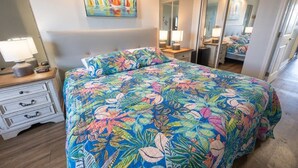 A bedroom with a floral bedspread, a bedside table with a lamp, a dresser, and a large mirror. - New at Caravelle Resort - Pools - Honeymoon Suite! (Myrtle Beach)