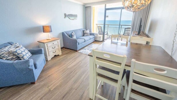 A living room with a sofa, a dining table with chairs, a lamp, and a view of the ocean through large windows. - New at Caravelle Resort - Pools - Honeymoon Suite! (Myrtle Beach)