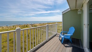 Um deck de madeira com guarda-corpo branco, dando vista para uma praia com guarda-sóis e cadeiras de descanso.
