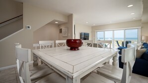 Uma sala de jantar espaçosa com uma mesa e cadeiras brancas, um arranjo central vermelho e vista para o oceano através de amplas janelas.