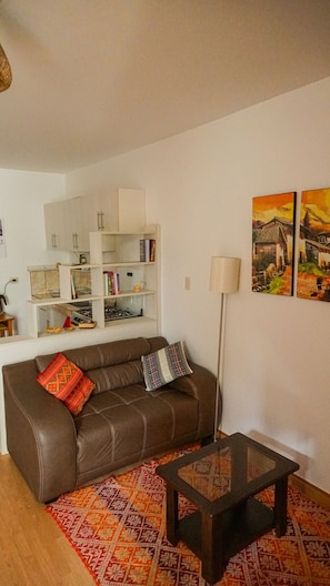 A living room with a brown leather sofa, a glass coffee table, a patterned rug, a white shelving unit with books and decorative items, and two landscape paintings on the wall.
