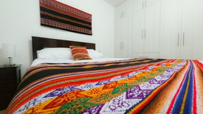 A bed with a colorful blanket, a white pillow, and a patterned pillow. There is a nightstand with a lamp and a wall-mounted painting above the bed.
