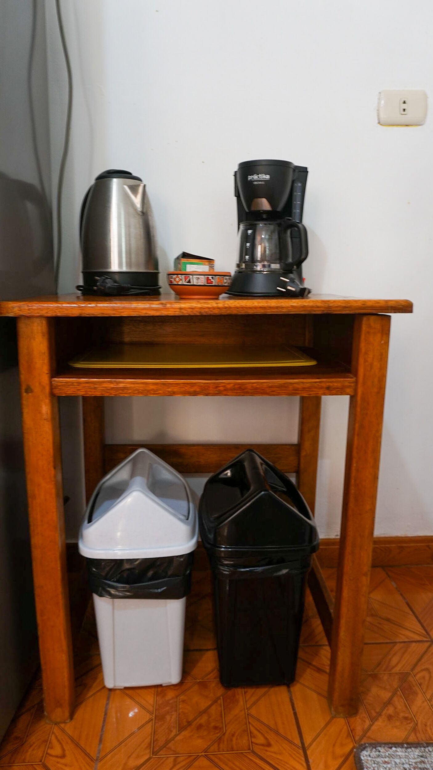 Ein Holztisch mit einem Wasserkocher, einer Kaffeemaschine und zwei Mülleimern darunter.