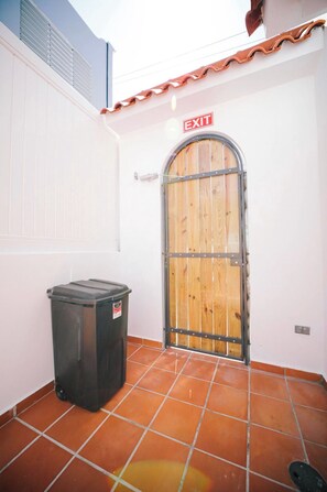 Una puerta de madera cerrada con marco de metal y un letrero que dice 'EXIT' arriba. Un bote de basura negro está colocado a la izquierda de la puerta.