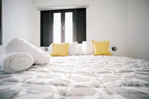 Un lit bien fait, avec des serviettes blanches pliées en rouleau et deux oreillers jaunes, une fenêtre avec des volets fermés et un mur blanc.