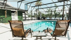 A poolside area with two wicker chairs and a small table, surrounded by a glass enclosure with a view of palm trees and a building in the background. - * Beautiful Lakehouse with Pool and Movie Theater* (Casselberry)