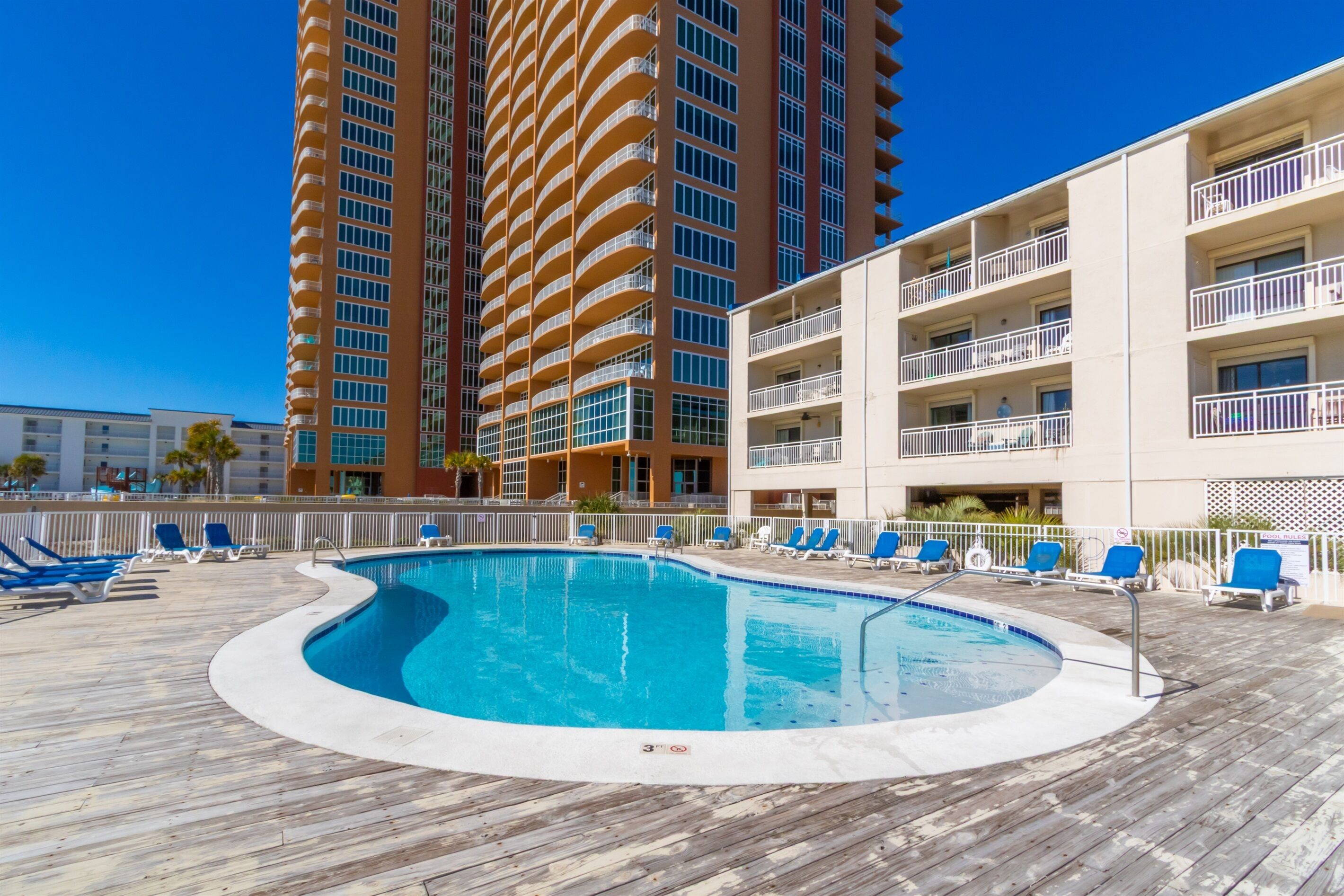 A pool area with blue lounge chairs and a pool, surrounded by a wooden deck and a building with multiple balconies.