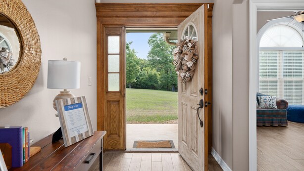 Una puerta de madera con vistas a un patio cubierto de césped, un espejo de mimbre, un letrero enmarcado y un banco con almohadones al fondo.