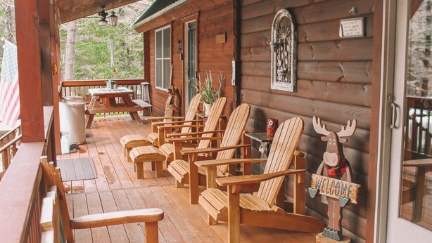 Eine Holzhütte mit Veranda, ausgestattet mit mehreren Adirondack-Stühlen, einem Schild mit einem Elchmotiv und einer Flagge.