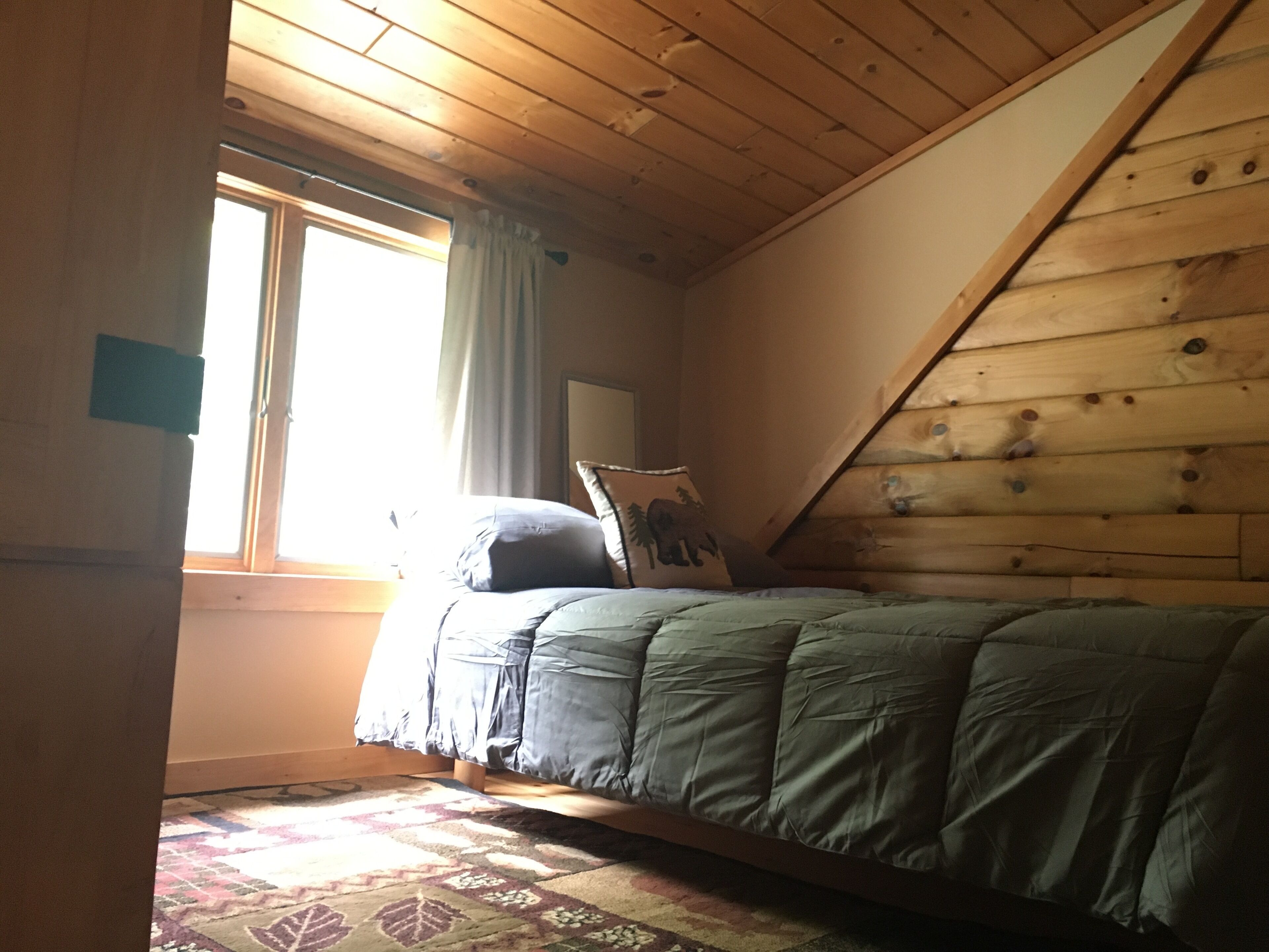 Un dormitorio con techo inclinado, una cama con colcha verde, una alfombra estampada y un ventanal con cortinas.
