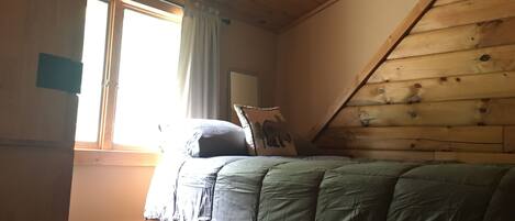 Un dormitorio con techo inclinado, una cama con colcha verde, una alfombra estampada y un ventanal con cortinas.