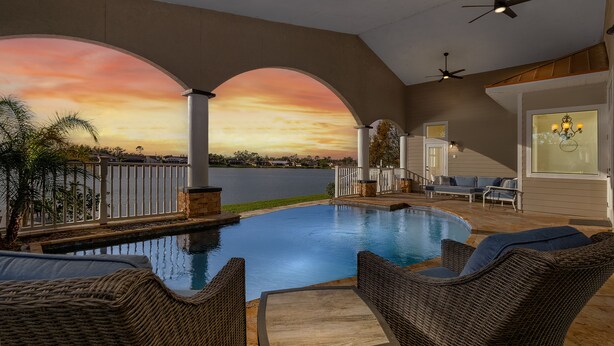 A pool area with wicker furniture and a view of a sunset over water.