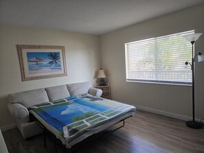 A bedroom with a bed, a nightstand, a lamp, a window with blinds, and a framed picture on the wall.