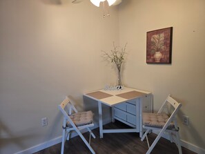 A small dining area with a white table and two chairs, a vase with branches, and a painting on the wall.