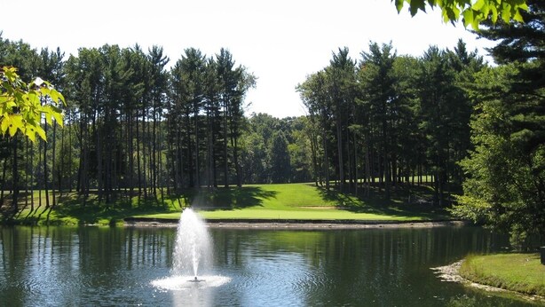 A tranquil pond with a fountain, surrounded by a well-maintained grassy area and tall trees. - The Pines of Apple Valley, 31 Beautiful Vacation Condos. (Howard)
