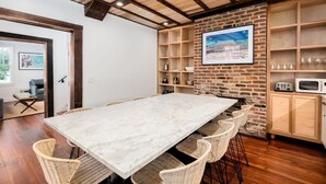 A spacious dining area with a marble table and wicker chairs, a brick wall with a framed picture, and wooden shelves with glasses and kitchen appliances. - Historic Boutique Home ‘Tuckertown’ (Charleston)