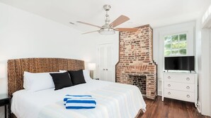 A bedroom with a bed, a brick fireplace, a ceiling fan, a TV, and a window. - Historic Boutique Home ‘Tuckertown’ (Charleston)