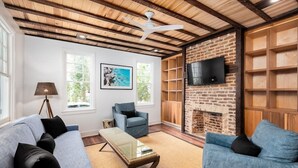 A living room with a blue sofa, a blue armchair, a coffee table, a fireplace, a television, and a ceiling fan. - Historic Boutique Home ‘Tuckertown’ (Charleston)