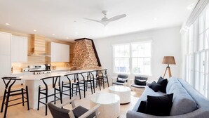 Una cocina moderna con pared de ladrillo, armarios blancos y una isla central con taburetes de bar. Hay un rincón comedor con un sofá y una lámpara.