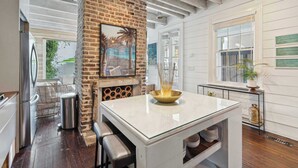 A modern kitchen with a brick accent wall, a glass-top island, and a view of the outdoor patio. - Private Pool & Historic Home - ‘Sugarsweet House’ (Charleston)