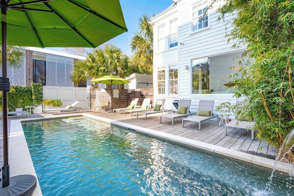 A swimming pool with lounge chairs and a green umbrella, surrounded by a wooden deck and a white building with large windows. - Private Pool & Historic Home - ‘Sugarsweet House’ (Charleston)