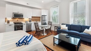 A modern hotel room with a bed, a sofa, a dining table with chairs, a kitchen area, and large windows with blinds. - Boutique Historic Home ‘The Cisco’ (Charleston)