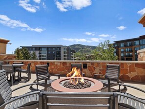 A rooftop patio with a fire pit surrounded by metal chairs, overlooking a modern building complex and a mountain range. - Sundial C407 | Ski-in/Ski-out | Village Views | Pools & Hot Tubs | Fitness Room | Ski Valet (Park City)