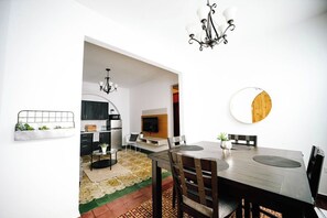 Un coin repas avec une table en bois, des chaises et un lustre. On aperçoit une cuisine à l’arrière-plan, équipée d’appareils électroménagers modernes et de meubles de rangement.