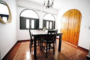 A dining area with a table, chairs, and a chandelier. There are two windows with shutters and a wooden door.
