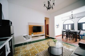 A modern living room with a flat-screen TV, a dining area with a table and chairs, and a kitchen area with a refrigerator.