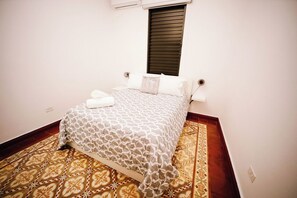 Une chambre à coucher avec un lit, une fenêtre avec des stores, un tapis à motifs et une prise électrique au mur.