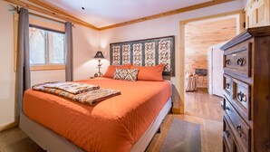 Un dormitorio con una cama grande, un ventilador de techo, una ventana con cortinas, una mesita de noche con lámpara y un aparador de madera.