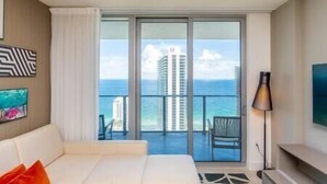 A hotel room with a large bed, a TV, a lamp, and a balcony with a view of the ocean and a tall building.