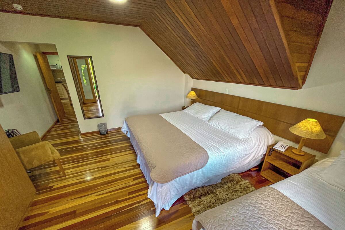 Habitación de hotel con piso de madera, una cama con ropa de cama blanca, una mesita de noche con lámpara y una pequeña mesa con teléfono.