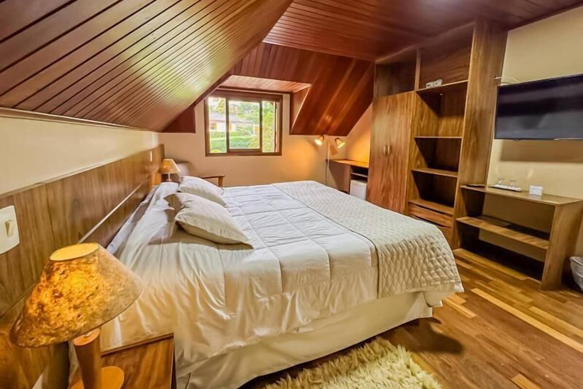Un dormitorio con techo inclinado, una cama con ropa de cama blanca, una lámpara, un televisor y estanterías de madera.