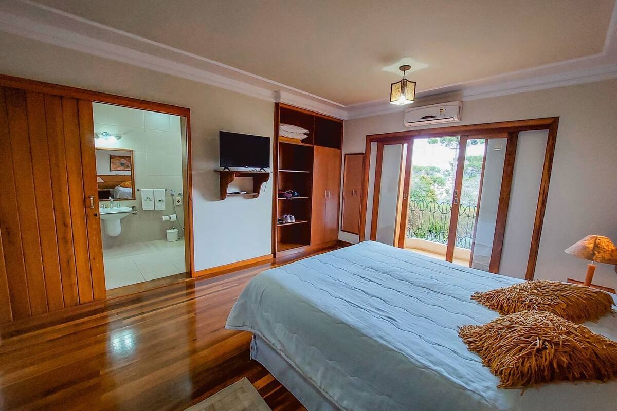 Un dormitorio con cama, suelo de madera, televisor, armario y baño visible a través de una puerta corredera.