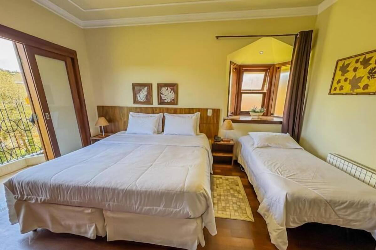 Habitación de hotel con una cama grande, dos camas más pequeñas, una ventana con cortinas y una planta en el alféizar.