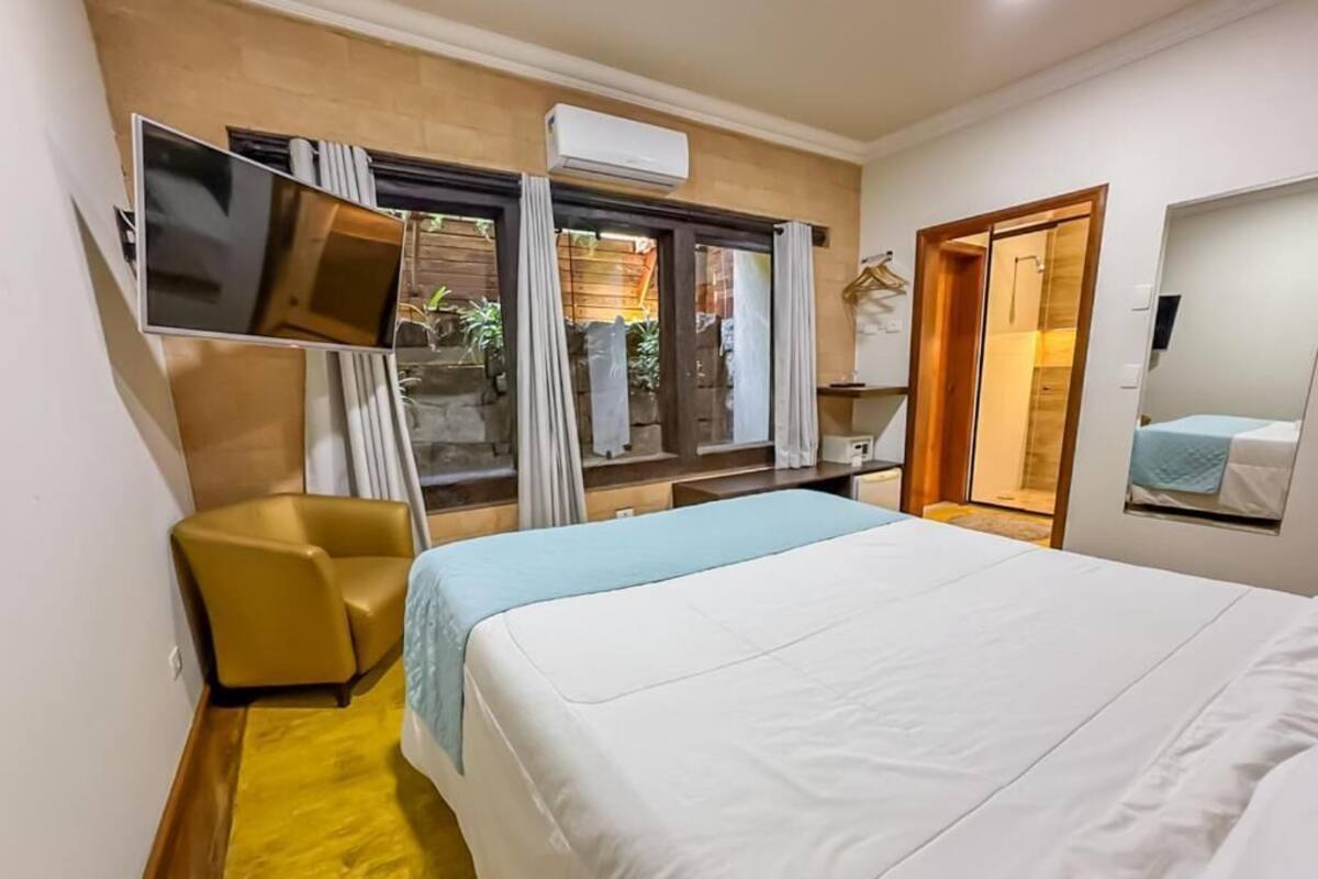 Una habitación de hotel con cama, televisión montada en la pared, una silla y una ventana con cortinas.