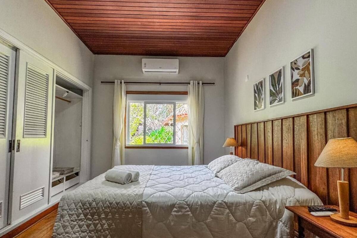 Un dormitorio con techo de madera, una cama con manta acolchada, una mesita de noche con lámpara y una ventana con cortinas.