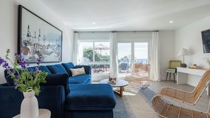 Un salon moderne avec un canapé bleu, un fauteuil en osier, une table basse et une grande fenêtre donnant sur la plage.