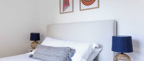 A neatly made bed with white linens, a gray throw blanket, and a blue lamp on a small round table beside it.