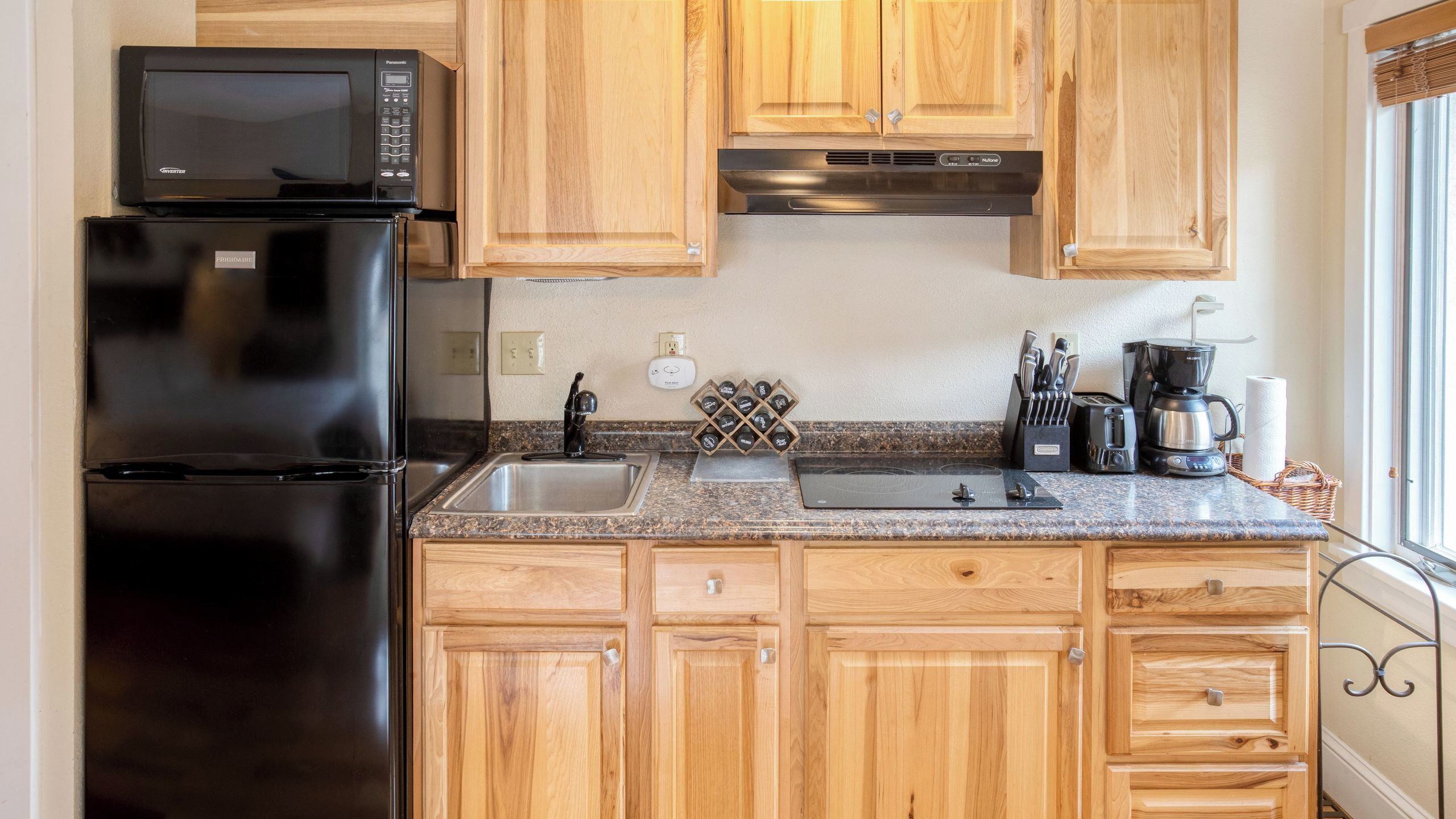 A kitchen with wooden cabinets, a black refrigerator, a microwave, a stainless steel range hood, a sink, a wine rack, and a coffee maker.