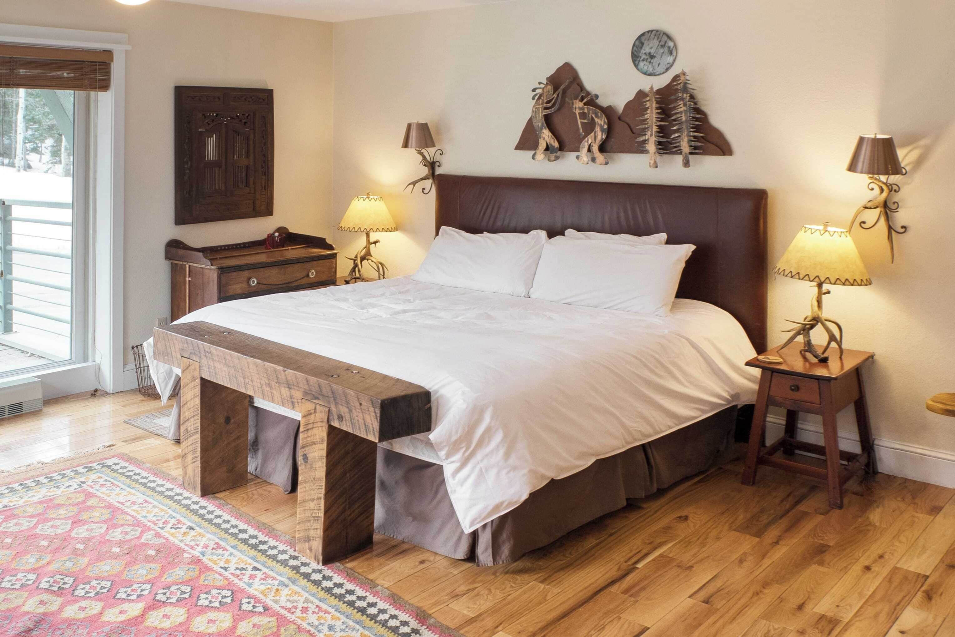 Une chambre à coucher avec un grand lit, une tête de lit en bois, deux tables de chevet avec des lampes, un banc au pied du lit, un tapis à motifs et une horloge murale.