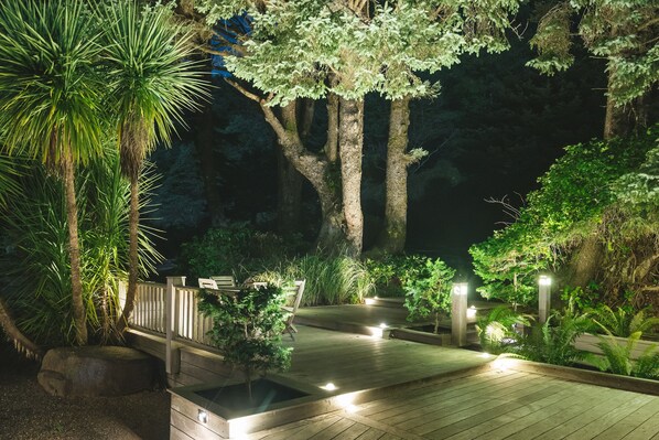 A wooden deck with benches, surrounded by lush greenery and trees, illuminated by soft lighting.