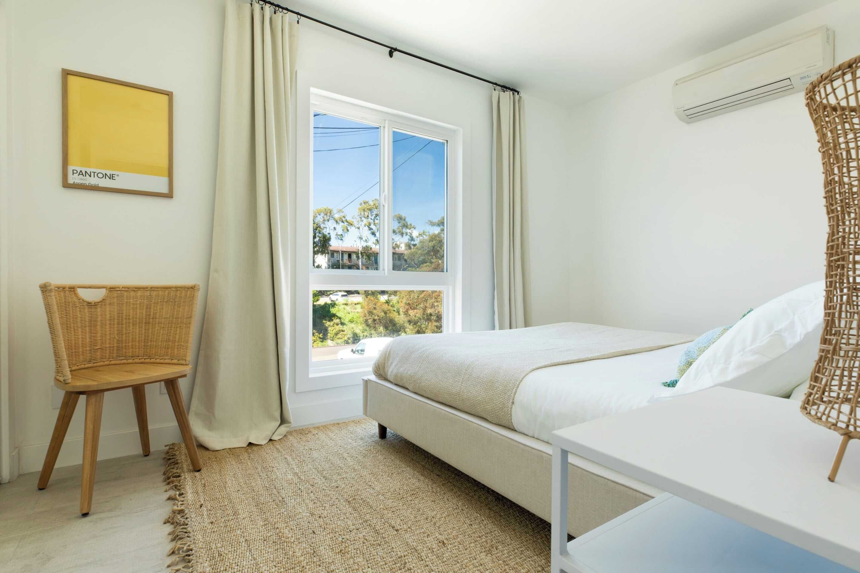 A bedroom with a bed, a chair, a window with curtains, and a wall-mounted air conditioner.